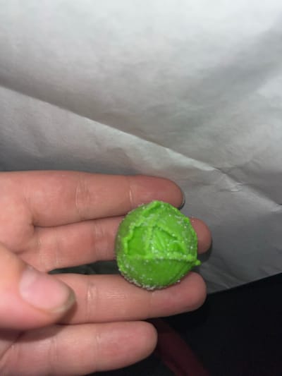 A close-up, slightly blurry shot shows a person's hand holding a bright green, ball-shaped candy. The candy has a textured surface that resembles cabbage leaves, and it appears to be dusted with fine white sugar crystals. The hand, with its fingers curled around the candy, is illuminated from above, creating subtle shadows. The background is a mix of white paper, possibly packaging, and a dark, out-of-focus area. The lighting suggests an indoor setting, likely at night or in a dimly lit room. There is no visible text in the image. The overall impression is that of someone holding a small, decorative candy.