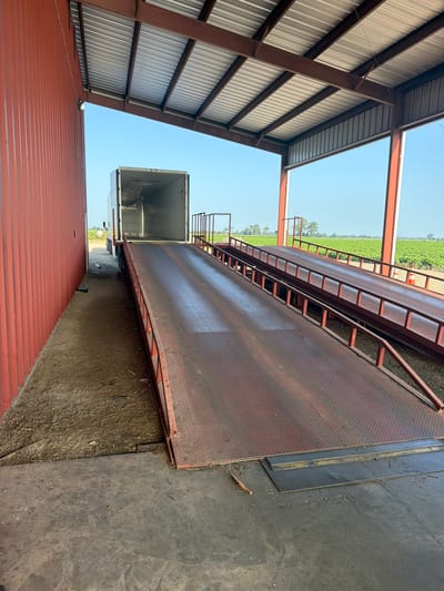 An outdoor loading dock area is depicted. A large, red-sided industrial building with a corrugated metal exterior is on the left. Its extended roof, supported by brown metal beams, covers the loading dock. Two red metal ramps with diamond plate surfaces and guardrails are positioned on the right, leading up to an open trailer of a white semi-truck. The truck is backed up to the building, with its trailer doors open. Beyond the loading dock, a green agricultural field with rows of crops stretches into the distance under a clear blue sky. Orange traffic cones are visible in the field. The ground in the foreground consists of concrete and gravel.