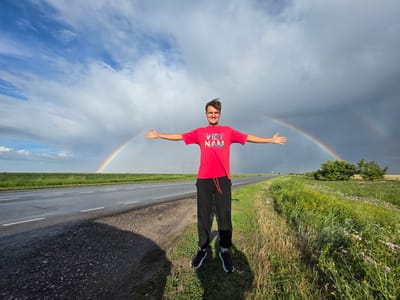 A young man stands in the middle of a rural landscape with his arms outstretched. He is wearing a red t-shirt that says "VIET NAM" and black pants. A double rainbow arches across the sky behind him, with dark storm clouds to the right and brighter, partly cloudy skies to the left. The man is standing on the side of a wet asphalt road that stretches into the distance. The road is bordered by a grassy field on the right and a green field on the left. In the distance, there are a few trees. The overall impression is one of a brief moment of beauty and peace amidst a dramatic weather event. This scene is not in Borodino, Russia, but rather appears to be in a flat, open landscape with a highway.
