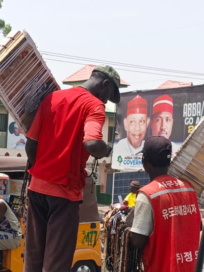 The image captures an outdoor urban scene in Hotoro, Nigeria, during daylight. Two dark-skinned males are prominently featured from behind in the foreground, each carrying a rectangular wooden drum on a shoulder. The individual on the left wears a camouflage cap, a red short-sleeved shirt over an orange undershirt, and dark trousers, with beaded necklaces around his neck. The individual on the right wears a black baseball cap and a red safety vest with white reflective strips and Korean text ("시루3동," "유도화감시자," and a partial "정") over a light-colored shirt, with multiple strands of beaded necklaces draped over his arm.
In the mid-ground, a large black political campaign banner displays portraits of two men in traditional white attire and red caps, identifying them as "ABBA GIDA GOVERNOR KANO STATE." Solar panels are visible beneath this banner. A partial view of a yellow tricycle, bearing the number "2477," is present on the left. Another individual wearing a yellow shirt and a brown hat is seated behind the right foreground figure, surrounded by numerous beaded items. The background includes light-colored buildings with reddish-brown tiled roofs and overhead wires against a bright, clear sky.