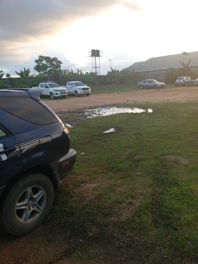 The image depicts a partly overcast outdoor scene, likely in Igrita, Nigeria, based on the prompt. A dark-colored SUV is partially visible on the left edge of the frame, its rear wheel and side panel are prominent. In the background, there are two white pickup trucks parked on a dirt patch. Further back, other vehicles are parked near a building with a long, slanted roof. A water tower is visible in the distance, standing above the tree line. The ground consists of a mix of dirt and grass, with puddles of water. The general ambiance suggests a rural or semi-rural setting.