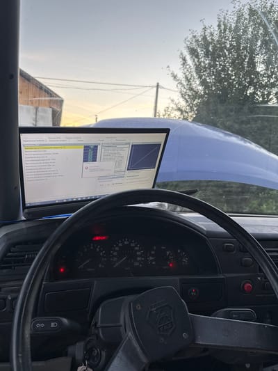 The image shows the interior of a vehicle, likely a car, with a laptop displaying diagnostic software open on the dashboard. The laptop screen shows various parameters related to engine diagnostics, including engine coolant temperature, crankshaft position, and throttle position. The dashboard features a steering wheel, instrument cluster with gauges for speed and RPM, and various indicator lights, one of which is red and labeled "STOP". The hood of the vehicle is open, revealing a blue engine bay. The scene appears to be outdoors, with a tree and a building visible through the windshield. The sky suggests it may be late afternoon or early evening. The image is taken from the driver's perspective. Based on metadata analysis, the image was geotagged in Krasnokamsk, Russia, suggesting the vehicle is located there. No people are visible in the image.
