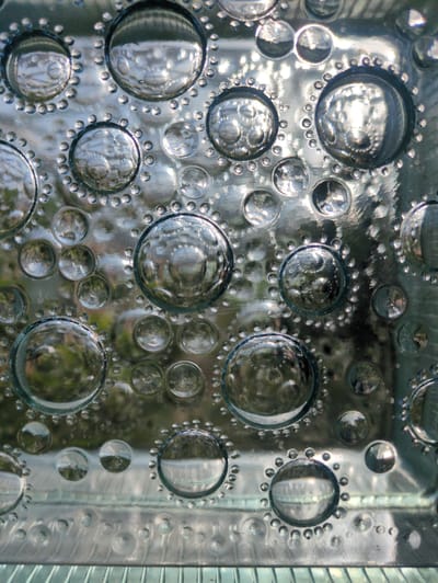 This media file displays a close-up view of a glass block with a textured surface. The glass block is comprised of numerous circular indentations of varying sizes, some of which are deep and contain reflective interiors, while others are shallower and appear as smaller bubbles. These indentations are arranged in a somewhat organized, yet irregular, pattern across the surface of the glass. The glass itself is transparent and shows reflections of an outdoor scene. Visible within the reflections are hints of green foliage, suggesting it is daytime and possibly a mild weather condition. The texture of the glass creates a distorted, fragmented view of the environment behind it. The overall impression is one of architectural detail or decorative element, possibly used in a building's facade or interior. No people, text, or specific activity are depicted. The location context of Velikovo, Russia, does not provide any directly visible cues within the image itself, but suggests the setting is within this geographical area.