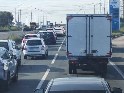 The image depicts a traffic jam on a highway in Vasilevka, Russia. The foreground shows the rear of a white vehicle, with a dark gray vehicle to its left. The road is lined with several cars and trucks, all stopped in traffic. In the background, a yellow tanker truck is visible, along with a gas station sign for "GAZPROM." The sky is clear and blue, and the overall lighting suggests it is daytime. The scene is indicative of typical highway congestion, with no unusual or dangerous activities apparent.