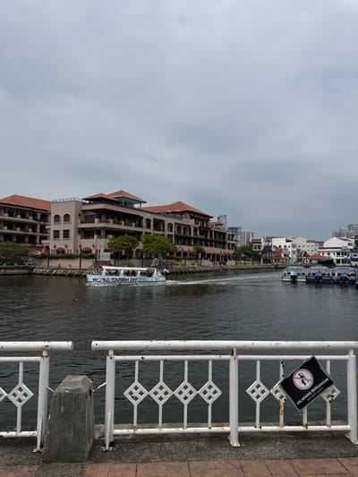 A waterway is lined with buildings and a white metal railing. The building on the left has tan walls and a red-tiled roof, while other buildings can be seen along the opposite bank. A white boat marked with "World Tourism Day 2025" traverses the waterway. A black and white sign with an image of crossed oars and text in Malay is affixed to the railing. The sky overhead is overcast.