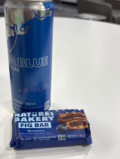 A blue Red Bull Juneberry Edition energy drink and a Nature's Bakery Blueberry Fig Bar sit on a white surface. The can is prominently displayed, showcasing the flavor and energy drink properties. The fig bar, also blueberry-flavored, highlights its whole grains, real fruit, and plant-based nature. The bar also notes it does not contain high fructose corn syrup and has a verified non-GMO logo. The nutritional information of the bar states it has 200 calories per pack.