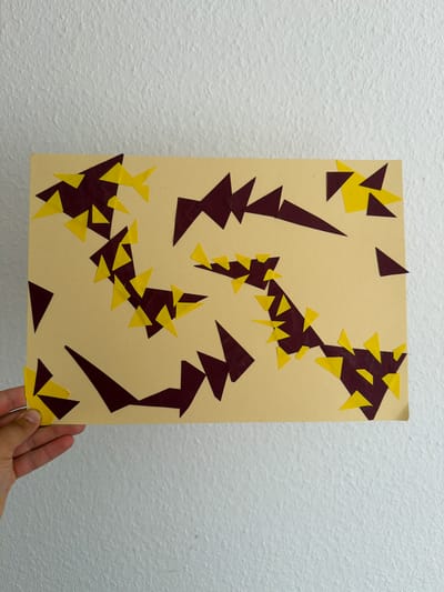 The image shows a close-up of a piece of abstract art made from paper cutouts. The art is affixed to a light beige rectangular sheet of paper and is displayed on a textured white wall. The cutout shapes are in two colors: a dark reddish-brown and a bright yellow. The shapes are primarily triangles and irregular polygons, arranged in a seemingly random but visually dynamic pattern across the beige background. Some of the cutouts are layered over each other.

A hand, likely belonging to the person holding the artwork, is visible at the bottom right corner of the frame, gently supporting the piece. The lighting appears to be neutral, suggesting an indoor setting, possibly an art studio or a home. There are no discernible people, animals, or specific objects that would indicate a narrative or scene, other than the artwork itself and the supporting hand. The background, the textured white wall, provides a neutral backdrop that emphasizes the artwork. There is no visible text in the image. The overall impression is of a simple, yet engaging, abstract composition.
