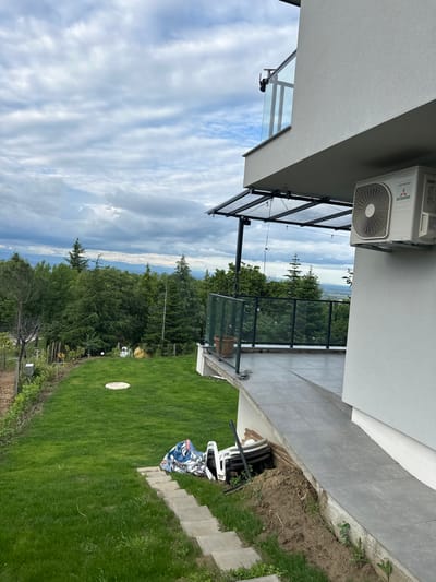 The image shows a scene in Brestnik, Bulgaria. The view from a modern building overlooks a lush green landscape with trees and hills in the distance, partially obscured by a cloudy sky. In the foreground, a tiled balcony with a glass railing extends from the building. Below the balcony, a well-maintained lawn slopes downwards. A small, circular white object is visible on the grass. Near the edge of the building, a partially obscured white object appears to be covered with a blue and white patterned sheet, possibly furniture. The building itself has a gray exterior and an external air conditioning unit.