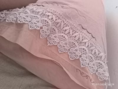 A close-up, overhead shot showcases the delicate white lace trim bordering a soft pink fabric, likely a pillowcase or bedding. The lace exhibits an intricate pattern of floral motifs and geometric shapes. Beneath the lace, the pink fabric is layered, creating subtle folds and shadows. Faint, decorative stitching in a darker thread can be seen on the pink fabric, forming swirling vine-like patterns. The overall impression is one of softness and understated elegance. The image is dated "17/07/2026 18:29". There is no indication of a specific location within the city of Şırnak, Turkey, in the provided image.