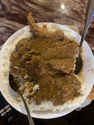 A close-up, overhead shot captures a generous serving of food in a white, round plate, resting on a brown and beige swirled table surface. The dish consists of white rice at the base, generously topped with a rich, dark brown, thick stew or sauce. Several pieces of meat, including one with a visible bone on the left side of the plate, are integrated into the stew. Two metal spoons are present; one is partially visible on the upper left, while the bowl of another spoon rests within the food on the lower right, suggesting a meal in progress or about to be eaten.

Adjacent to the plate, a small, light-colored cloth or napkin is tucked underneath. On the bottom right of the frame, a black remote control is partially visible, displaying text such as "JVC", "Netflix", "Live", "Menu", and "Back", indicating a domestic setting. The scene depicts a home-cooked meal, typical of the provided location context of Lakka, Sierra Leone. The lighting suggests an indoor environment.