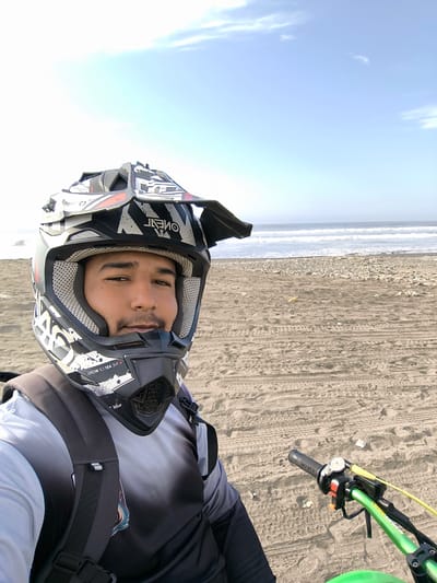 A selfie of a man wearing a full-face O'Neal motocross helmet with a visor and carrying a backpack. He is on the beach, seated on a green dirt bike. The background is a sandy beach with tire tracks, leading to the ocean and a slightly hazy sky. The scene is likely Huanchaco, Peru.