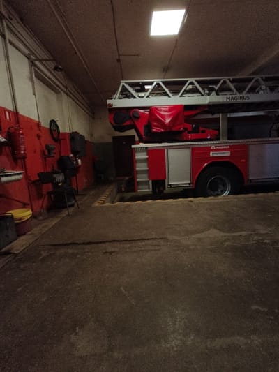 A bright, square-shaped fluorescent light fixture is visible on the ceiling of a dimly lit room. Below the light, a red and silver fire truck with the brand "MAGIRUS" emblazoned on its side is parked. The truck has a large, extendable ladder mechanism on top, covered in places by a red tarp. To the left of the truck, a red wall is adorned with various items, including a fire extinguisher, a metal sink basin, and a black stool. The floor appears to be a concrete or asphalt surface, showing signs of wear and tear. Yellow and black markings are visible on the floor in front of the fire truck, suggesting a designated parking or operational area. The overall scene suggests an indoor environment, likely a fire station or garage.