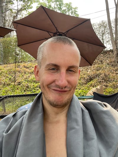 A man with short, light gray hair, a nose ring, and stubble is positioned in front of a brown patio umbrella. He is wearing a gray garment, and his chest is exposed. The setting appears to be outdoors, with vegetation and another umbrella visible in the background. The photo was taken in Tambon Nam Phrae, Thailand.