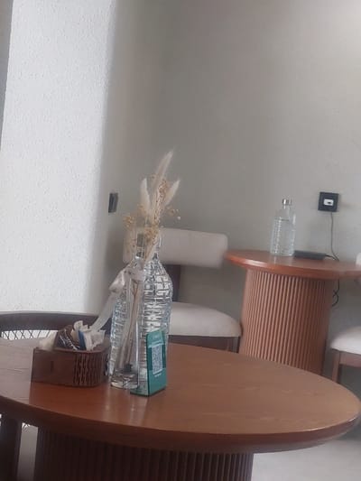 The image depicts a close-up view of a wooden table in what appears to be a cafe or restaurant setting in Kyzylorda, Kazakhstan. On the table, there is a clear glass vase containing dried floral arrangements and a ribbon tied around its neck. Next to the vase is a small wooden box filled with sugar packets and other sachets. A QR code on a teal stand is also visible on the table, likely for menu or payment purposes. In the background, a bottle of water sits on a fluted, orange-colored side table. The walls are textured and light-colored. Two light-colored chairs with wooden frames are partially visible in the background. The overall impression is of a quiet, interior scene within a commercial establishment.
