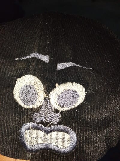 The image shows a close-up of an embroidered design on a black fabric. The design depicts a cartoonish face with large, circular eyes outlined in light gray and filled with dark gray. Above the eyes are two thin, curved eyebrows. The nose is a small, triangular shape. The mouth is open, revealing embroidered teeth in a light gray color, outlined with dark gray. The embroidery is detailed, with visible stitching patterns. The fabric appears to be textured, possibly corduroy or a similar material. The lighting in the image is dim, creating shadows and highlighting the texture of the fabric and embroidery. There is no indication of the specific location in Rumuomasi, Nigeria, or any context beyond the embroidered image itself.