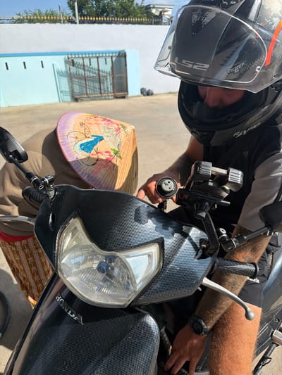 An eye-level shot captures a person wearing a black motorcycle helmet and a t-shirt, positioned next to a black Honda scooter. The helmet features "LS2" on its clear visor and "CREEP" and "RDO" visible on its surface. The person's face is obscured by the helmet, but tattooed forearms, tanned skin, and a watch on the left wrist are visible. The t-shirt shows a partial "ADIDAS" logo.

The scooter, adorned with a checkered, carbon fiber-like pattern on its front fairing, displays the "HONDA" emblem near its headlight. A motorcycle phone mount is attached to the handlebars. The person's right hand holds a small cylindrical object with three pins, possibly a key or charging component, near the phone mount, while the left hand rests on the handlebar grip.

A traditional Vietnamese conical hat (nón lá), painted with a scene of a person on a bicycle, rests on a surface to the right of the scooter's front wheel. In the background, a light blue building with a barred window and multiple vents is visible under a clear, bright sky, suggesting daytime. Green foliage from trees can be seen further in the distance on the left. The scene evokes an outdoor setting in Ninh Chu Ward, Vietnam, with strong sunlight illuminating the subjects.