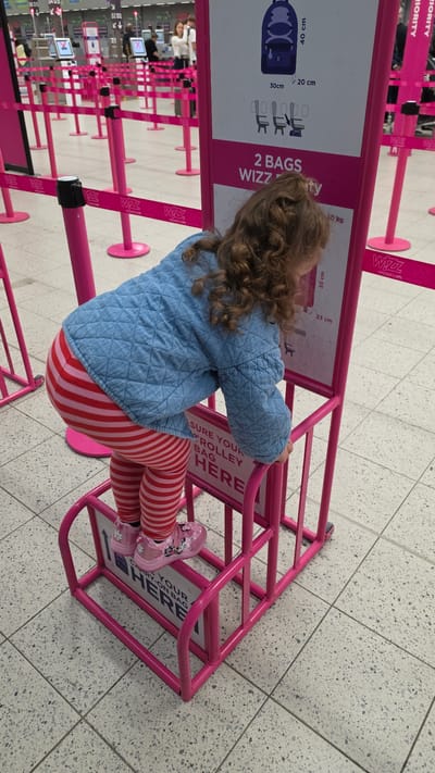 A young child is depicted from behind, standing next to a pink Wizz Air baggage sizer in an airport terminal in Luton, United Kingdom. The child has curly brown hair and is wearing a light blue quilted jacket, red and white striped leggings, and pink shoes featuring a cartoon character, possibly Minnie Mouse.
The prominent pink metal baggage sizer is designed to check luggage dimensions, with visible text indicating "SURE YOUR TROLLEY BAG HERE" and "CARRY-ON BAG HERE". An attached sign displays baggage size limits, including "40 cm", "20 cm", "30 cm" for a backpack icon, and "55 cm", "23 cm" for another bag, along with "10 kg". The sign also states "2 BAGS WIZZ".
The floor is made of light-colored speckled tiles. In the background, a queue area is formed by numerous pink stanchions, each bearing the "Wizz" logo and "wizzair.com" URL. Other travelers and airport kiosks/monitors are faintly visible further back, suggesting a busy airport environment. Partial text like "PRIORITY" is seen on a banner in the upper left. The image captures a moment of a child in an airport setting, likely at a check-in or bag drop-off point for Wizz Air.