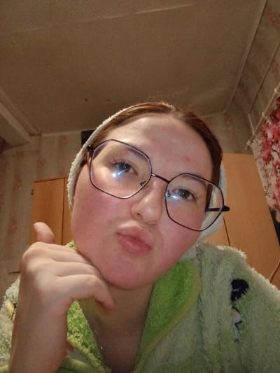 A close-up, upward-angle indoor photograph depicts a female subject from the upper torso, framed against an interior background. The subject has reddish-brown hair, partially covered by a white, towel-like headband, and wears oversized, dark-rimmed, geometric-shaped prescription glasses. Her face exhibits a generalized reddening, particularly on the cheeks and forehead, and her lips are pursed forward. She is attired in a textured, light green garment, consistent with a robe or a fuzzy top, which features darker green accents or patterns. Her right hand (viewer's left) is positioned under her chin, with the thumb extended vertically in a gesture. Bright light reflections are prominently visible on the lenses of her glasses. The background consists of a light-colored ceiling. To the left of the subject, a wall covered with patterned wallpaper is visible. To the right, a dark, vertical piece of furniture or wall section is partially obscured by a curtain or fabric displaying a red and white floral pattern. The precise location in the city of Старый Шагирт, Russia, cannot be determined from the visual evidence presented in the image.