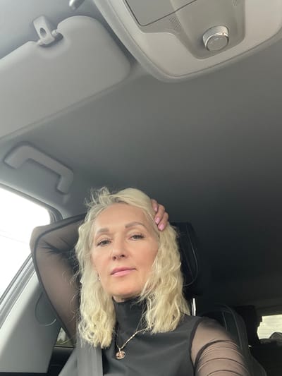 In the interior of a vehicle, a blonde woman with curly hair is seated, resting her right arm on the headrest and her hand behind her head. She is wearing a black, high-necked garment with sheer, long sleeves and a gold necklace with a pendant. She has on light-colored eye makeup, lip gloss, and a nose ring. The vehicle's sun visor and a part of the ceiling are visible in the upper portion of the image. The scene is set in the city of Ul'yanovka, Russia.