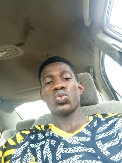 A young man with dark hair and skin puckers his lips in a kissy face. He is seated in a car, evidenced by the car ceiling, a headrest, and a window behind him. He wears a patterned black and white shirt with yellow accents. The lighting is diffused, and the overall image has a casual, selfie-style composition.