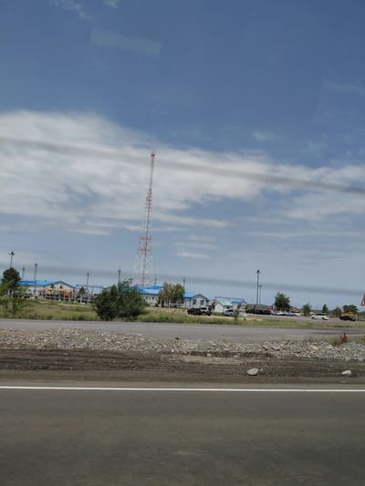 A cell tower stands tall in the background of this image, which appears to have been taken from a moving vehicle. In front of the tower are several buildings with blue roofs. There are also some trees and a road visible. The sky is cloudy. The scene looks like it could be in Qazaqstan, Kazakhstan.