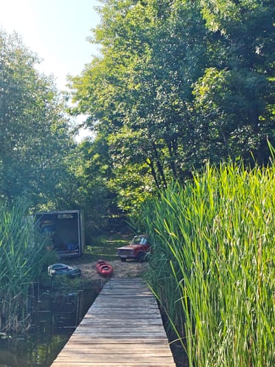 The image depicts a sunlit scene at the edge of a body of water, likely a lake or river, in Horodyshche, Ukraine. A wooden pier extends from the right into the water. Tall, green reeds line the bank on both sides of the pier. To the left of the pier, on the shore, is an inflatable boat, dark in color. Next to the inflatable boat is a red kayak. Behind the boats, a small, open-fronted shed or trailer is visible, containing various items. Further inland, a red, vintage car is parked on a dirt path. The background is dominated by lush green trees, suggesting a natural, possibly wooded, environment. The sky is bright and clear. There are no people visible in the image.