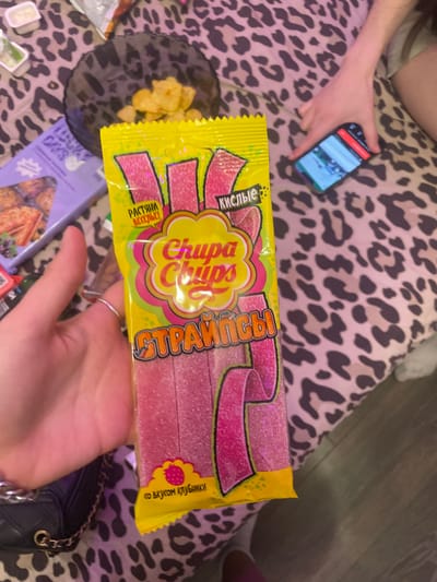 In the city of Zmeinyi, Russia, a person is holding a yellow package of "Chupa Chups" sour straws. The package is pink and features the Chupa Chups logo. The straws themselves are also pink and appear to be sprinkled with sugar. In the background, a hand is visible holding a smartphone, and a leopard-print blanket covers the surface. There are also some chips in a black bowl and a purple box in the background.