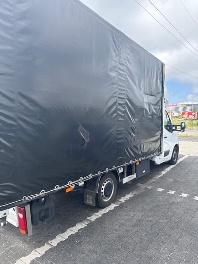A white truck with a large black tarp covering its cargo area is parked on a paved lot. The truck is positioned at an angle, with the front facing away from the camera. The tarp is draped over the cargo area, creating a dark, opaque surface that obscures whatever is inside. Some of the tarp is wrinkled and appears to be secured with ropes and metal grommets.
In the background, a building with a checkered facade is visible, along with a red sign that reads "Media Markt". Another sign above it indicates "850m" suggesting distance. The sky above is partly cloudy with a few visible power lines running horizontally. The ground is paved with interlocking concrete blocks and white dashed lines marking parking spaces. The scene appears to be an outdoor commercial area, likely a parking lot in Zgorzelec, Poland, given the context. There are no people visible in the image.