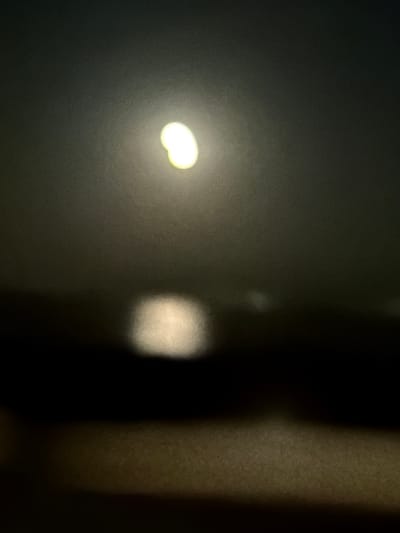 The image depicts a dark scene with a blurred bright light source in the upper portion of the frame, possibly the moon or a street light. Below this, a darker horizontal band suggests a landscape or body of water, with another blurred bright area in the lower center. The overall atmosphere is indistinct due to the blurriness. No specific location in Noratus, Armenia can be identified from this image. There are no people, objects, or actions discernible.