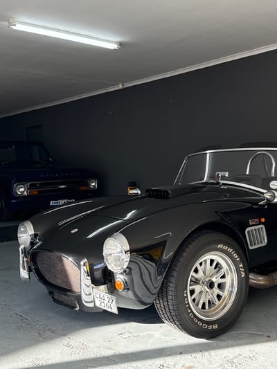 The image shows a black AC Cobra sports car parked in a garage in Blouberg, South Africa. The car is positioned in the foreground, facing slightly to the left. Behind it, a dark blue Chevrolet pickup truck is partially visible. The garage has a grey concrete floor and dark grey walls. A fluorescent light fixture is mounted on the ceiling. The AC Cobra features a prominent hood scoop, round headlights, and a wire mesh grille. Its license plate reads "CAA 27 2164". The tire on the right side of the Cobra is a BFGoodrich Radial T/A. Sunlight casts shadows on the floor from the cars.