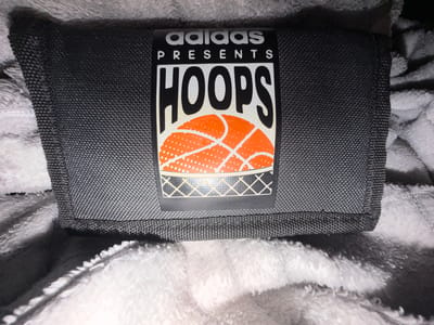 The image shows a close-up, overhead view of a dark grey fabric object, possibly a wallet or pouch, with an "Adidas Presents Hoops" logo. The logo features the Adidas name in white, followed by "PRESENTS" in smaller white text, and "HOOPS" in large black letters. Below "HOOPS" is a graphic of an orange basketball with white dots and black lines, and a white net pattern. The object is placed on a soft, white, fluffy surface. The image does not contain any information about the city of Dorsten, Germany.