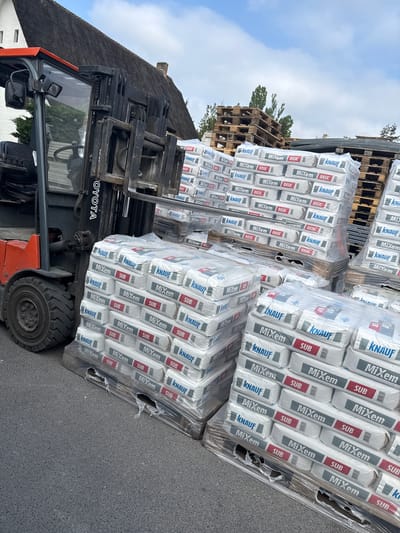 An outdoor scene captures a forklift positioned next to multiple pallets laden with construction material bags. The forklift, an orange and black Toyota model, has its forks extended, supporting a large stack of wrapped bags. In the background, additional pallets stacked with similar bags are visible, along with empty wooden pallets piled high. Further back, trees and the edge of a building with a traditional thatched roof can be seen. The sky is partly cloudy, indicating daytime.

The activity appears to involve the movement or storage of these construction materials, with the forklift ready to transport the loaded pallet. The scene suggests a building supply yard, a construction site, or a delivery point for construction materials.

Visible text includes "TOYOTA" on the forklift. On the white bags, brand names "KNAUF" (in blue) and "MiXem" (in black) are prominent. Some bags are labeled "BASIC" and others "SUB," both in red text against a white or grey background.

The image was captured in Boortmeerbeek, Belgium.
