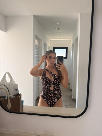 A young woman stands in a white, modern hallway, taking a selfie in a mirror. She is wearing a leopard-print one-piece swimsuit with cut-out details. Her hair is pulled back in a ponytail, and she is touching her ear. To her left, on a surface in front of the mirror, are a brown designer handbag and a white plastic shopping bag. The hallway has white walls, a tiled floor, and recessed ceiling lights. In the reflection of the mirror, another white door and a dark-framed window are visible. The overall setting suggests an indoor, possibly hotel, environment in Trachoni, Cyprus.
