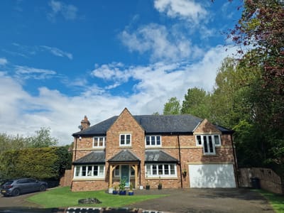 The image depicts a two-story brick house under a partly cloudy blue sky. The house has a dark gray roof and white-framed windows. The front door is light blue, sheltered by a wooden-framed porch. Several potted plants are arranged in front of the porch. A white garage door is visible on the right side of the house. A grey car is parked in the driveway to the left of the house, partially obscured by a tall green hedge. Mature trees are present on the right and in the background. The house sits on a green lawn, separated from the road by a strip of grass and a small stone barrier. The location is likely a residential area in the city of Durham, United Kingdom.