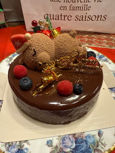 This image depicts a festive chocolate cake situated indoors within the city of Kawasaki, Japan. The round chocolate cake features a glossy dark ganache topping. It is adorned with two textured brown bear figures, one larger and one smaller, positioned centrally; the larger bear wears a red and white Santa hat with a snowflake pattern. Other decorations include a gold-colored "Happy Christmas" script, a small gold-colored leaping reindeer, two red raspberries, two dark blue blueberries, and a cluster of miniature red and silver jingle bells with a green artificial pine accent. The cake rests on a white cardboard base, which is placed on a white porcelain plate with a blue and purple floral rose pattern. A red tablecloth or placemat underlies the plate. In the background, a light-colored paper gift bag is visible, bearing French text that reads "une nouvelle vie en famille quatre saisons." Behind the bag, a red surface with a gold geometric pattern is partially visible. No individuals are present in the scene.