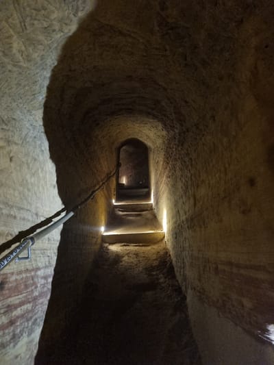 The image depicts a tunnel or cave-like structure. The interior surfaces are rough and appear to be made of stone or rock. At the end of the tunnel, there are openings that let in light. A pipe or pole runs along the bottom of the tunnel toward the viewer. The location is in Homburg, Germany.