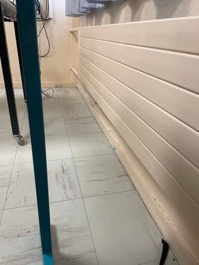 The image depicts an interior scene, specifically a corner of a room in Hérouville-Saint-Clair, France. The floor is covered with light-toned rectangular tiles exhibiting mottled grey and brown patterns. The walls are painted a uniform light beige or cream color.
Along the right wall, a cream-colored horizontal panel radiator is mounted, featuring multiple parallel fins. Below the radiator, the wall's base shows evidence of chipping and debris accumulation. A thin, cream-colored pipe is visible running horizontally along the wall above the radiator, bending downwards on the left side, and another horizontal pipe is partially visible below the radiator, obscured by the wall damage.
In the foreground, the lower sections of what appear to be two table or desk legs are present. On the left, a dark-colored, possibly black, square-section leg is visible, equipped with a grey caster wheel at its base. To its right, a teal-colored vertical support leg extends upwards from the floor.
Behind the teal leg and against the left wall, a tangle of black and grey electrical cables descends from an unseen point higher up. A white rectangular surface, possibly an electrical outlet or junction box, is mounted on the wall where these cables are present. On the floor directly below these cables, a reddish-brown circular mark or stain is discernable. At the extreme bottom-right corner of the image, a small black, curved object rests against the wall, consistent with a tool handle or debris. No individuals are present in the scene.
