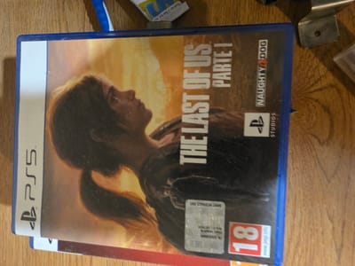 The image shows a PlayStation 5 game case for "The Last of Us Part I" lying on a wooden surface. The game case is oriented vertically, with the front cover facing the viewer. The cover art features a close-up of a woman's face in profile, with a blurred background of warm, sunset-like colors. The game's title and "Part I" are prominently displayed in white lettering. Below the title, the "Naughty Dog" logo is visible, along with the "PlayStation Studios" logo. The "PS5" logo is also visible on the left side of the case. To the right of the game case, a silver metal bracket with a hole is partially visible. A small portion of another game case, with a red edge, is seen beneath the primary game case. The scene appears to be indoors, possibly on a table or shelf, given the wooden texture of the surface. There is no identifiable indication of the city of Brindisi, Italy, within the image itself.