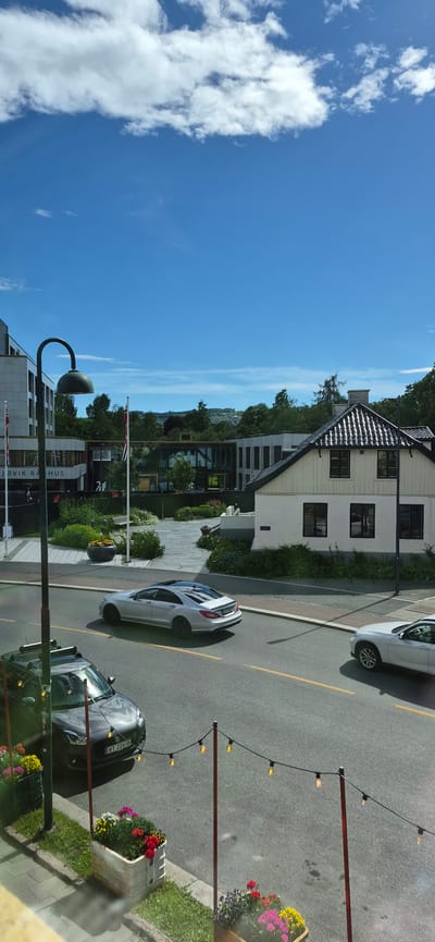 This is an outdoor, eye-level, medium shot taken from a slightly elevated perspective, depicting a street scene on a sunny day in Gjøvik, Norway. The image is oriented vertically, with the sky occupying the top left portion and the street running diagonally from the bottom right to the upper left.

The sky is a vibrant blue, with scattered white clouds. To the right of the sky is a row of buildings. The most prominent building in the middle ground is a two-story structure with a dark, tiled roof and light-colored siding. It has several windows. To the left of this building, and closer to the foreground, is a modern building with large glass windows, suggesting it might be an office or commercial space. The text "GJØVIK RA" is partially visible on this building.

On the street, there are two cars. A silver car is parked on the right side of the road. Further down the street, a darker colored car is also parked. The road is asphalt with a yellow line marking the lanes. String lights with exposed bulbs are visible in the foreground, strung between two posts, adding a decorative element. There are planters with colorful flowers, including red, pink, and yellow blooms, along the sidewalk. Green grass and trees are also visible, interspersed with the buildings and sidewalk.

The sunlight suggests it is daytime, likely late morning or early afternoon. The clear blue sky and sunlight indicate fair weather. There are no people visible in the frame, but the presence of cars, buildings, and streetlights implies an urban or town setting. The Norwegian flag is visible on a flagpole near one of the buildings. The overall mood is serene and bright, capturing a typical day in this Norwegian town.