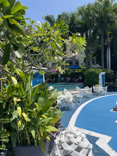 In Bang Sao Thong Subdistrict, Thailand, a resort pool area is depicted on a sunny day. Lush green foliage, including plumeria and palm trees, frames the scene. A bright blue pool with a tiled edge is visible, adjacent to a paved area painted in contrasting blue and white. Scattered white metal tables and chairs are set up on the paved area, some occupied by lounge chairs and people relaxing. A signpost with directional arrows is positioned near the pool. In the foreground, a large planter holds more greenery. Several individuals are seen lounging by the pool, with one person standing near the water. The architecture of the resort buildings is partially visible in the background.