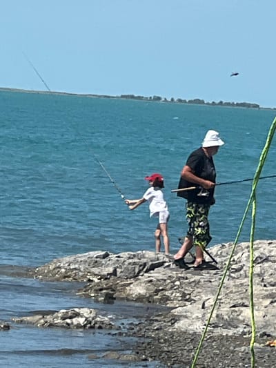 A man and a child are fishing on a rocky shore. The man, wearing a black t-shirt, white hat, and patterned shorts, stands to the right, holding a fishing rod. The child, in a white shirt and red hat, stands to the left, also with a fishing rod. They are by the side of a body of water, with a distant shoreline visible. The sky is clear and blue.
