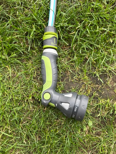 A garden hose nozzle, attached to a hose, lies on a patch of green grass with some bare soil. The nozzle is primarily gray and green, with various spray settings labeled on it. The scene appears to be outdoors, likely in a residential area. There are no people or animals visible in the image. The location is not identifiable from the image itself.