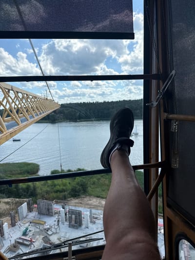 This media file is a photograph taken from inside a crane cab. The perspective is from a high vantage point, looking down and out through a large window. A person's leg, wearing a dark sneaker, is prominently featured in the foreground, resting on what appears to be a metal beam or ledge within the cab. The leg and foot are positioned towards the right side of the frame.
Through the window, a vast expanse of water dominates the middle ground, with a treeline visible on the far bank. The water's surface is calm, reflecting the sky. In the distance, a large yellow lattice structure, likely part of another construction crane or bridge, extends towards the water. The sky is filled with dramatic, fluffy white clouds against a bright blue backdrop.
To the right of the water, a construction site is visible. Several modular buildings or structures are laid out on a concrete surface, surrounded by building materials and equipment. The ground appears to be a dirt or gravel area, with some greenery interspersed.
The overall scene suggests a daytime setting, possibly late morning or early afternoon, given the bright lighting and shadows. The weather appears to be clear with some cloud cover. The location is identified as Mikołajki, Poland. The image conveys a sense of height and perhaps a moment of pause or rest for the crane operator. There is no visible text in the image.