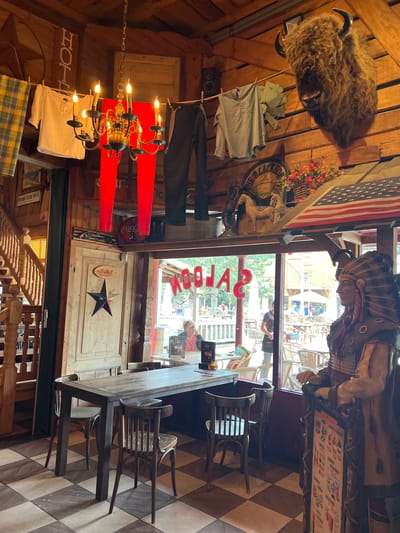 The image shows the interior of a rustic-themed establishment, possibly a restaurant or bar, located in Collendoorn, Netherlands. The walls and ceiling are constructed from wood, creating a warm and country-style atmosphere. In the foreground, a large wooden table with chairs is set on a checkered floor. To the right, a life-sized statue of a Native American chief stands prominently. Above the table, a chandelier with multiple candle-shaped lights hangs from the ceiling. Laundry, including a white shirt, red pants, and dark trousers, is hung on a clothesline across the upper part of the room. Mounted on the wall is a taxidermied bison head, adding to the wild west theme. Behind the table, a large window looks out onto an outdoor seating area with tables and umbrellas, and further beyond, buildings typical of a village or tourist attraction. On the window, the word "SALOON" is written in large, red lettering. The overall impression is of a themed establishment aiming for a nostalgic or adventurous ambiance.