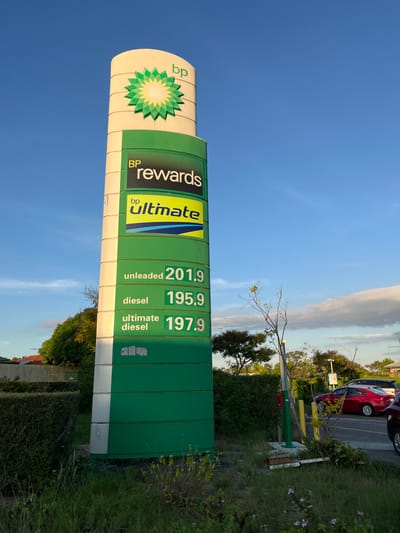 A green and white BP fuel station sign is standing upright in a suburban setting.  It advertises BP Rewards and BP Ultimate, with the current fuel prices: Unleaded - 201.9, Diesel - 195.9, and Ultimate Diesel - 197.9. The sign is in front of a hedge of greenery. On the right side of the sign is a small, white and green sign partially visible. A red sedan is parked in the background. There is a green pole to the right of the sign, a yellow sign on the ground, and a green post partially visible behind the sedan. The location of the sign, based on the provided coordinates, is a BP fuel station at 198B Pacific Hwy,  Gordon, NSW 2072, Australia.
