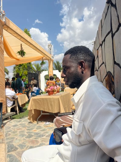 The image depicts an outdoor social gathering, consistent with a tropical climate, with an unspecified location within Kumasi, Ghana, as requested. In the immediate foreground, positioned to the right, a Black male is seated in profile, facing left. He wears a white, textured, collared shirt with a visible left chest pocket and white pants. His dark, short hair is closely cropped, and he has a short beard. He holds a dark smartphone in his left hand.
The background reveals an event setting under a large, light orange/peach fabric canopy supported by metal scaffolding. Decorative greenery hangs from the canopy structure. Beneath the canopy and extending into the middle ground, multiple rectangular tables are present, covered with matching light orange/peach tablecloths. One table near the center features a pot with pink floral arrangements. Numerous individuals, primarily adults of Black ethnicity, are gathered around these tables. They are seen standing, sitting on various chairs (including white plastic ones), and socializing. Some attendees wear vibrant, patterned clothing, while others are in more subdued attire, including some in green uniforms or outfits.
The ground is paved with irregular stone slabs in shades of grey and brown, transitioning to green grass further back. Beyond the event setup, tropical vegetation, including palm trees, is visible. A high wall constructed from large, irregularly shaped stones (brown, grey, white) borders the right side of the scene. The sky is bright blue with scattered white cumulus clouds, indicating a clear, sunny day. No specific landmarks or identifiers within the image indicate a precise location within Kumasi.