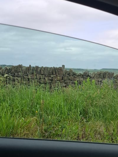 The image displays a rural landscape viewed through a car window. In the foreground, tall green grass and some wildflowers are visible. Behind the grass is a dry stone wall constructed from dark, weathered stones. Beyond the wall, the landscape stretches out with rolling hills, and several wind turbines can be seen in the distance. The sky is overcast with a pale grey hue. The location is identified as Barnsley, United Kingdom.