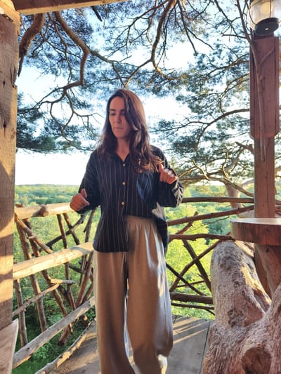 A woman stands on a wooden structure, possibly a treehouse or balcony, in Онохино, Russia. She has long, dark hair and is wearing a dark, pinstriped button-down shirt and light-colored sweatpants. Her hands are raised, and she appears to be gesturing or posing for a photo. The background shows a canopy of pine trees and a lush green forest. The lighting suggests it is late afternoon or early evening. The wooden structure features rustic construction with thick, rough-hewn logs and branches. A vintage-style lantern is visible on a wooden post to the right of the woman.