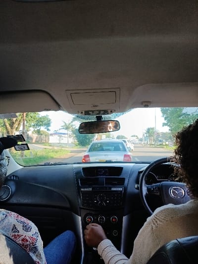 The image depicts an interior view from a vehicle in Harare, Zimbabwe, looking forward through the windshield during daylight hours. A driver with dark, curly hair, wearing a light-colored top, is positioned on the right, with their right hand grasping a manual gear stick. The vehicle's dashboard is black, featuring a multi-function steering wheel with a Mazda logo and a central console displaying infotainment and climate controls. A front passenger is partially visible on the left, wearing blue trousers and a patterned upper garment. The rearview mirror, centrally located, reflects the upper face and curly hair of an additional individual. Outside the vehicle, a two-lane road extends forward, with a silver sedan directly in front. Other vehicles are visible further down the road. The immediate roadside environment consists of green vegetation, various trees including palm trees, and obscured buildings consistent with an urban or suburban setting.