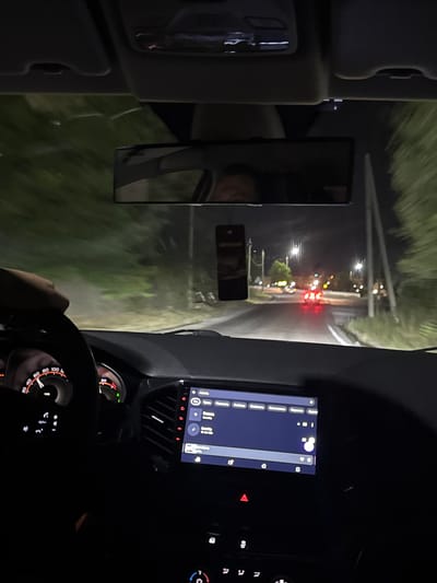 A first-person perspective shot from inside a car at night shows a hand gripping the steering wheel, with the dashboard illuminated. The car is moving on a road lined with trees, and there are streetlights and other cars visible in the distance. The car's infotainment screen is on, displaying song titles. The image is taken from inside a vehicle traveling on a paved road at night, likely on the outskirts of Chinarkent, Uzbekistan, given the style of the road and vegetation. The driver's hand is visible on the steering wheel, and the car's dashboard, including the speedometer and infotainment screen, is illuminated. The infotainment screen shows a list of music tracks. The road ahead is visible, with other vehicles' taillights illuminated and streetlights lining the sides of the road. The surrounding environment appears to be a mix of trees and vegetation, suggesting a semi-rural or suburban setting. The overall impression is that of a nighttime drive through an area with some artificial lighting and natural surroundings.